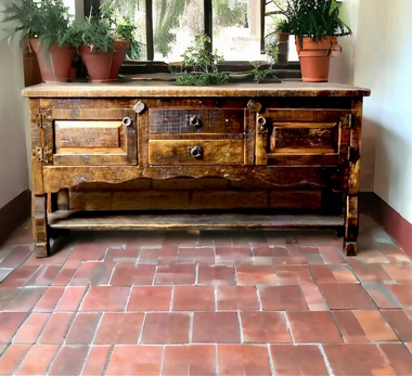 Rustic wood sideboard with iron hardware and carved drawers beneath a window of potted plants