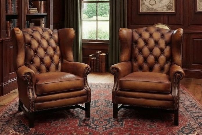 Pair of tufted tan leather wingback chairs with nailhead trim on a patterned rug in a wood-paneled library